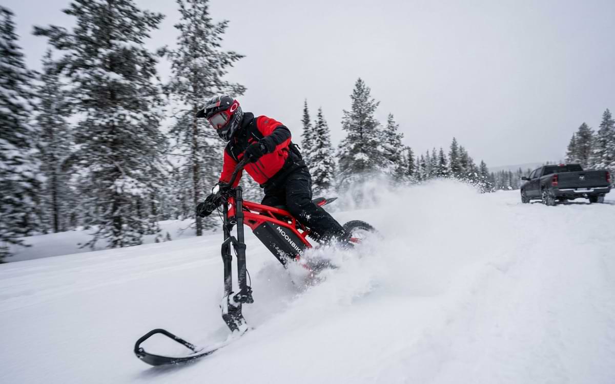 Rider in winter gear carving through fresh powder on a red Moonbike electric snow bike with a transport truck visible in the background.